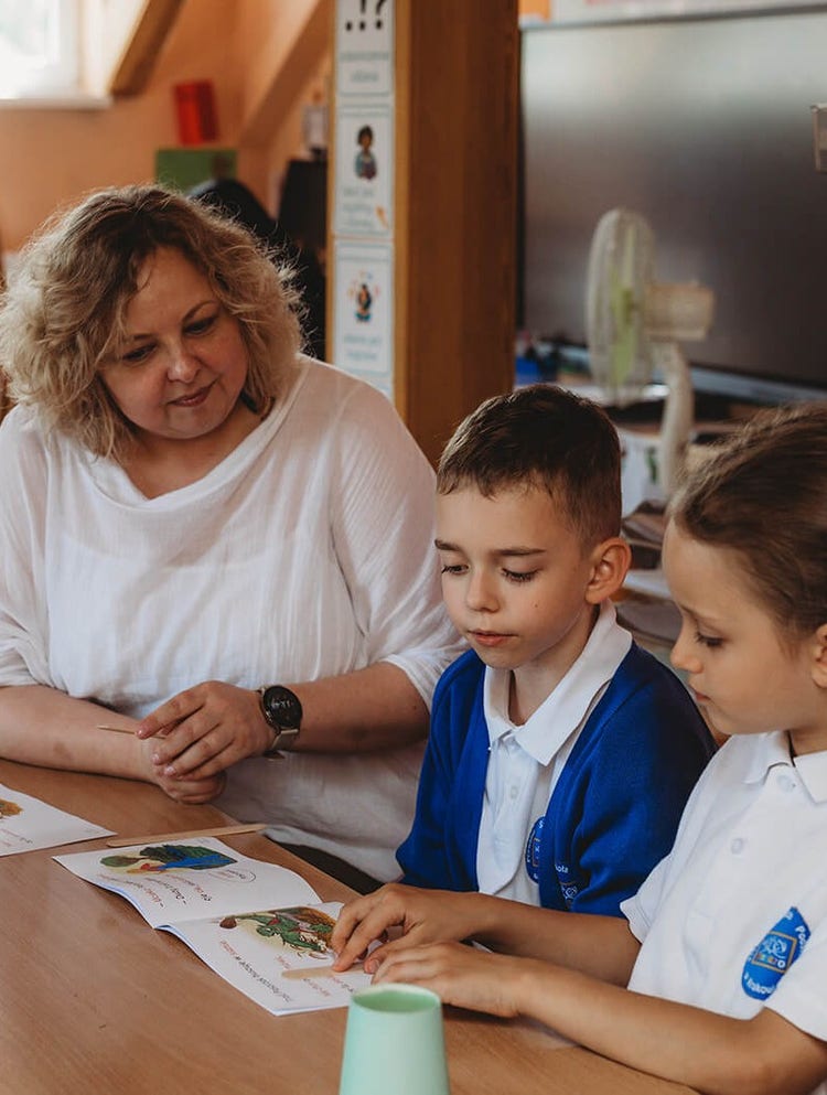 Dzieci z nauczycielką przy stole, czytające książeczkę, nauczycielka po prawej stronie ubrana w białą koszulkę, nauczycielka ma kręcone, jasne, średniej długości włosy, jest uśmiechnięta, przypatruję się co robią dzieci, dzieci po jej prawej stronie czytają książeczkę, pokazują drewnianym, jasnym, małym patyczkiem konkretne słowa, czytają, chłopiec po prawej stronie nauczycielki ma krótkie brązowe włosy, jest ubrany w jasne, białe polo z krótkim rękawem, niebieski sweter kardigan z logo prywatnej szkoły podstawowej, dziewczynka po jego prawej stronie jest bokiem, jest ubrana w białe polo z krótkim rękawem z logo szkoły podstawowej, dzieci są uśmiechnięte i skupione na zadaniu, w tle są inne dzieci siedzące na dywanie, są zajęte innymi aktywnościami, jasna, przestronna, nowoczesna sala z tablicami do nauki, jasne ciepłe kolory